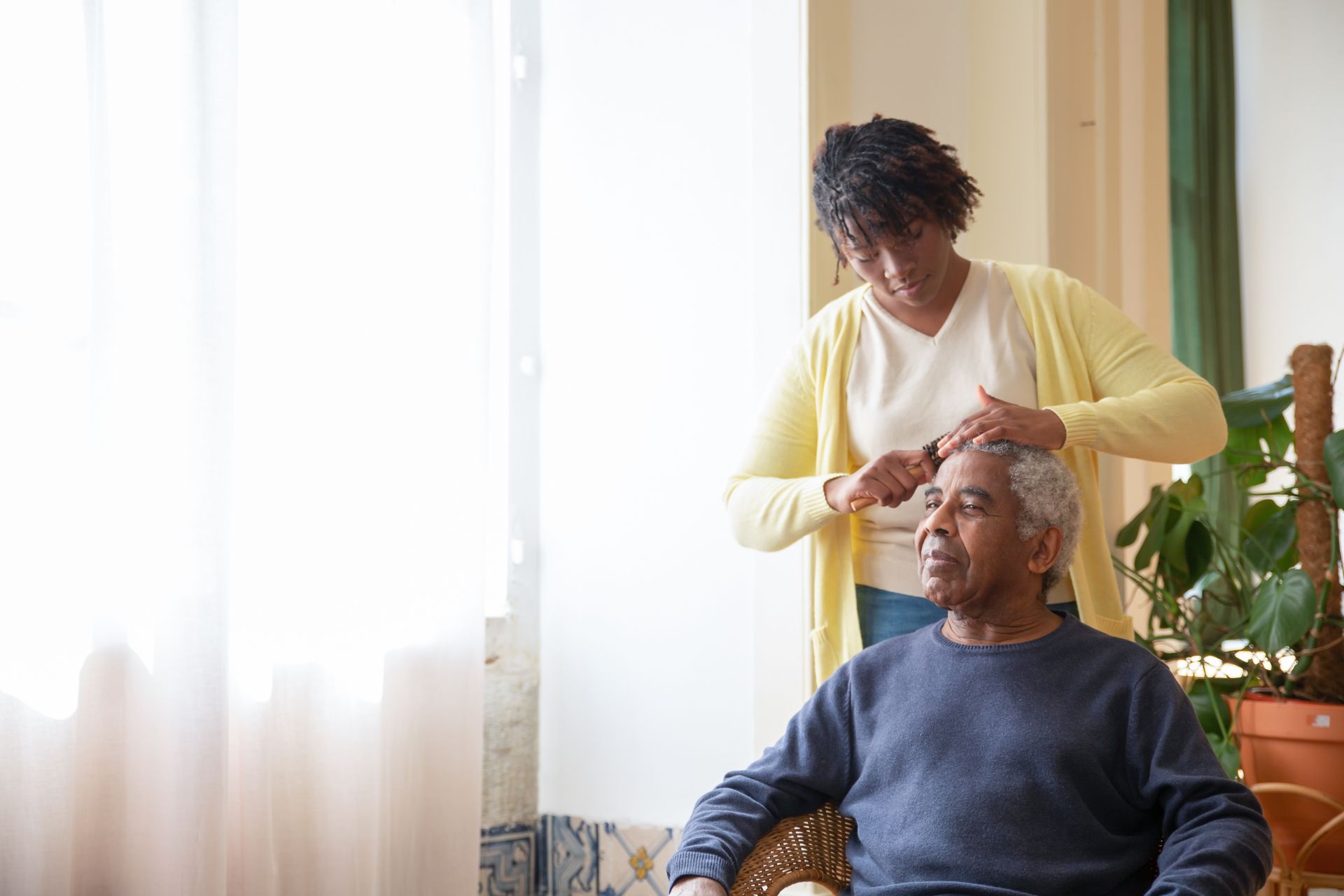 A caregiver gently combing an older man's hair by a sunlit window at home